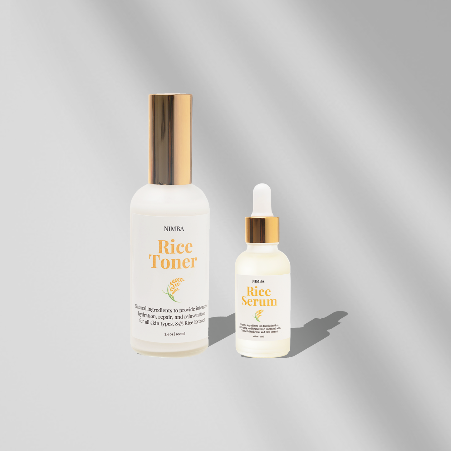 NIMBA Rice Toner and Rice Serum on a soft silver-gray background. The Rice Toner comes in a tall frosted bottle with a shiny gold cap, and the Rice Serum is in a smaller dropper bottle with a matching gold cap. Both labels highlight the rice-based skincare benefits for hydration and skin rejuvenation