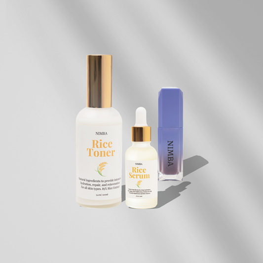 NIMBA Rice Toner and Rice Serum on a soft silver-gray background. The Rice Toner comes in a tall frosted bottle with a shiny gold cap, and the Rice Serum is in a smaller dropper bottle with a matching gold cap. Both labels highlight the rice-based skincare benefits for hydration and skin rejuvenation